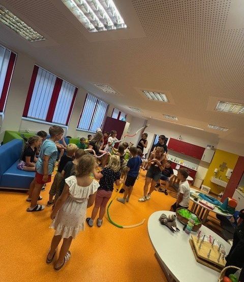 Eine Gruppe von Kindern spielt in einem hell beleuchteten Raum. Einige stehen und andere sitzen auf einer blauen Couch. Ein Kuchen und einige Spielzeuge stehen auf einem Tisch.