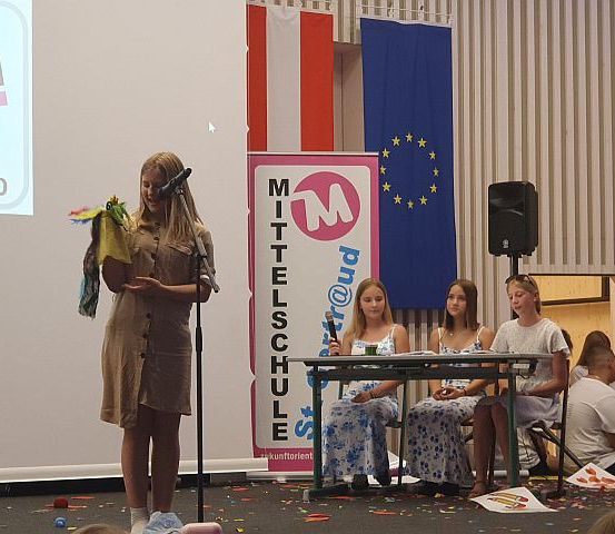 Eine junge Frau steht auf der Bühne mit einem Mikrofon, hält eine Puppe und spricht zu einem Publikum. Hinter ihr sitzen drei Mädchen an einem Tisch, und eine Projektionsleinwand ist an der Wand. Fahnen von Österreich und der Europäischen Union hängen darüber.