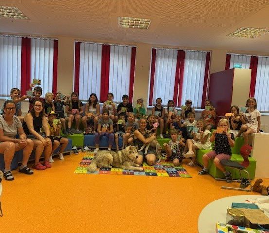 Eine Gruppe von Kindern und Erwachsenen versammelt sich in einem Raum mit einem Hund. Einige sitzen auf einer Couch, andere auf dem Boden. Sie halten Karten und Spielzeug. Eine Frau hält einen Hund, während sie kniet.