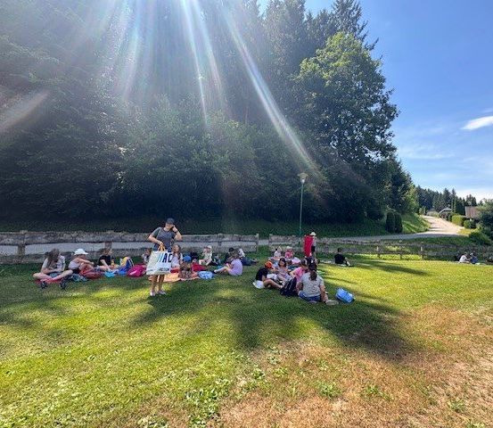 Eine Gruppe von Kindern und Erwachsenen sitzt auf einem Grasfeld, wahrscheinlich in einem Park. Sie sind an einem sonnigen Tag. Einige von ihnen tragen Hüte und Brillen. Eine Frau hält eine weiße Tasche mit dem Text 'AC'. Es gibt Bäume und Büsche in der Umgebung.