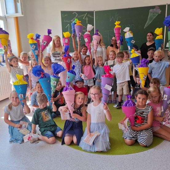 Eine Gruppe von Kindern und Erwachsenen in einem Klassenzimmer hält farbenfrohe Geschenkkegel und posiert lächelnd für ein Foto. Ein Tafel mit Zeichnungen ist im Hintergrund zu sehen.