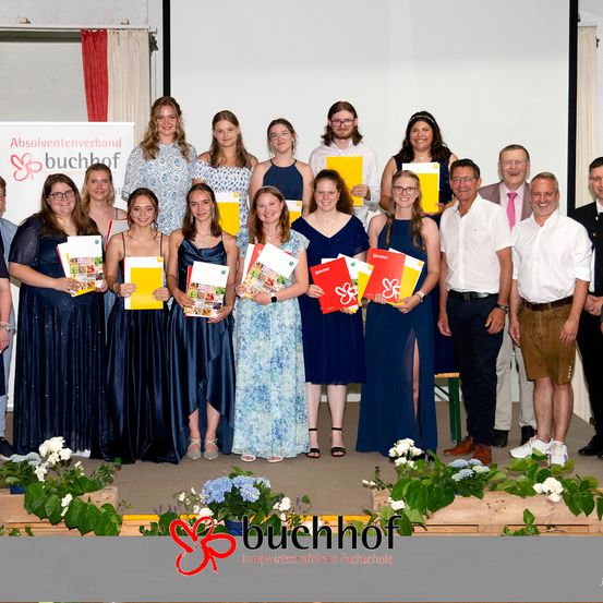 Eine Gruppe von Menschen in blauen Kleidern und weißen Hemden posiert für ein Foto auf einer Bühne und hält Urkunden in den Händen. Dahinter zeigt eine weiße Wand ein Banner mit Logo und Text.