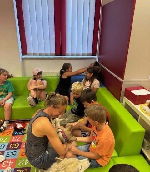 Eine Gruppe von Kindern sitzt auf grünen Sofas und interagiert mit Hunden in einem Raum. Eine Frau hält einen Hund, während ein Junge ihn streichelt. Ein Mädchen sitzt auf dem Sofa mit einem Plüschtier. Eine Mikrowelle steht auf dem Schreibtisch. Eine Aufbewahrungsbox ist neben dem Sofa.