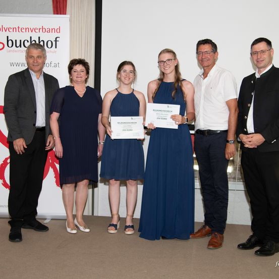 Drei Frauen und zwei Männer stehen vor einem Hintergrund mit einem Logo. Sie lächeln und halten Urkunden. Die Frauen tragen Kleider und Brillen. Die Männer tragen Anzüge. Die Frauen halten Urkunden mit der Aufschrift Bildungsscheck.