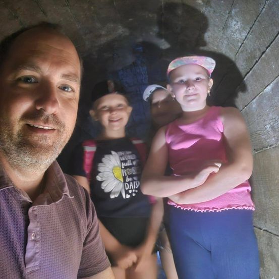 Ein Mann in einem braunen Hemd steht in einem Tunnel mit zwei Kindern, einem Mädchen in Pink und einem Jungen in Schwarz, beide lächeln.