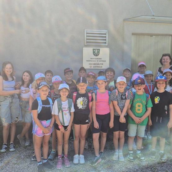 Eine Gruppe von Kindern und zwei Erwachsenen steht vor einem Gebäude. Einige der Kinder tragen Hüte und Rucksäcke. Das Schild lautet 'Marktgemeinde Frantschach-St.Gertraud'.