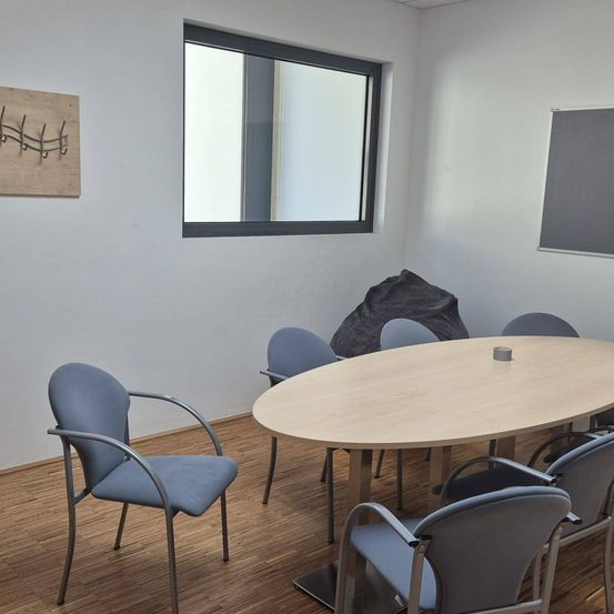Ein leerer Besprechungsraum mit einem runden Holztisch und blauen Stühlen. Ein grauer Sitzsack steht in der Nähe des Fensters.