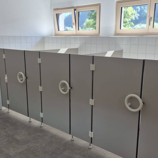 Ein Badezimmer mit grauen Kabinen, weißen gefliesten Wänden und zwei großen Fenstern mit Blick auf Bäume draußen.