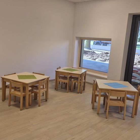 Ein leerer Raum mit Holzböden und weißen Wänden. Es gibt drei Holztische mit grünen und blauen Tischsets. Der Raum hat ein Glasfenster mit Blick auf einen Außenbereich.