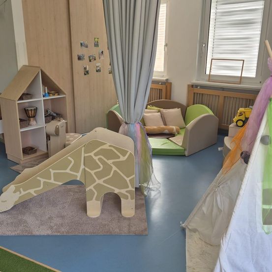 Ein Kinderspielzimmer mit einem hölzernen Giraffenrutsche, einem kleinen Haus auf Regalen, einem grünen Sofa mit Kissen und einem Zelt in der Ecke.
