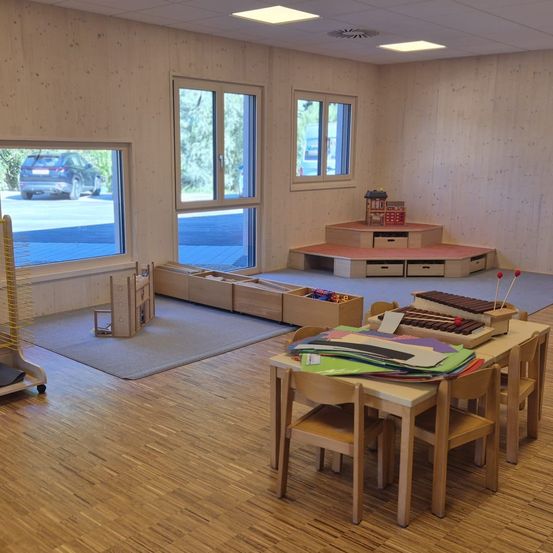 Ein leerer Raum mit Holzwänden und -böden. Ein Tisch mit verschiedenen Gegenständen steht in der Mitte. Ein Teppich liegt auf dem Boden. Ein Regal mit Gegenständen ist an der Wand. Ein Auto ist durch das Fenster sichtbar.