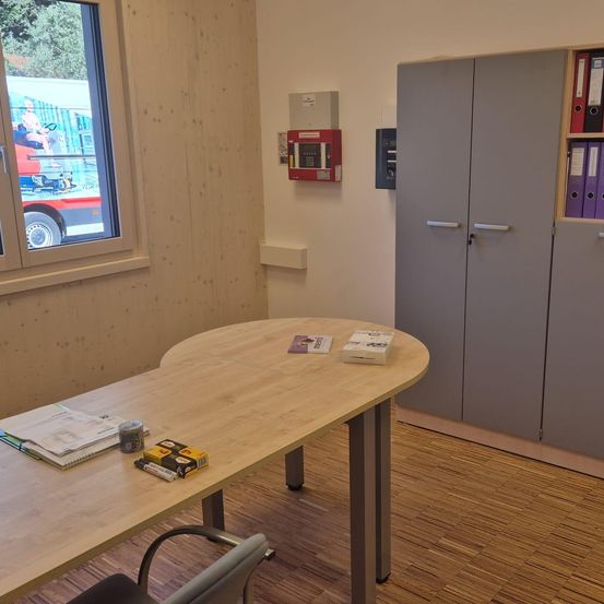Ein Büro mit einem Holztisch hat Bücher, Marker und andere Gegenstände. Ein Feuerlöscher ist an der Wand, und ein Feueralarm ist darüber. Ein Fenster zeigt einen Blick auf ein Fahrzeug draußen.