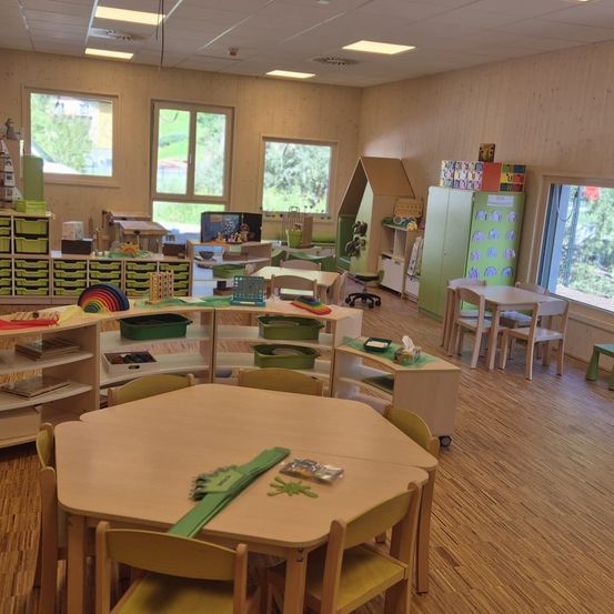 Ein Klassenzimmer mit Holzböden und -wänden, gefüllt mit Lernspielzeug, Büchern und bunten Regalen. Verschiedene Tische und Stühle sind für Kinderaktivitäten angeordnet. Fenster lassen natürliches Licht eindringen.