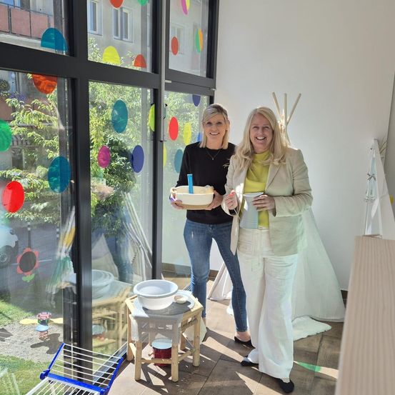 Zwei Frauen stehen in einem Raum mit bunten Glasfenstern. Eine hält einen Tablett mit Schüsseln, und die andere hält einen Becher. Ein kleiner Tisch mit Schüsseln und einem roten Eimer steht hinter ihnen. Eine zeltähnliche Struktur befindet sich rechts, und ein Auto ist durch das Fenster sichtbar.