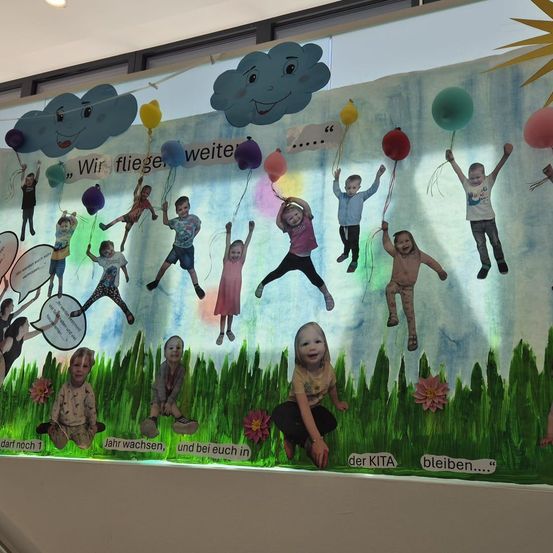 Eine farbenfrohe Ausstellung zeigt Kinder mit Luftballons, mit Text 'Wir fliegen, weit... Jahr wachsen, und bei euch in der KITA bleiben...'. Die Szene umfasst Wolken, Blumen und ein saftiges Feld.