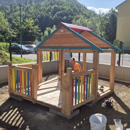 Ein Mann mit Hut arbeitet an einem hölzernen Spielhaus in einem eingezäunten Hof mit grünen Bäumen und geparkten Autos im Hintergrund.