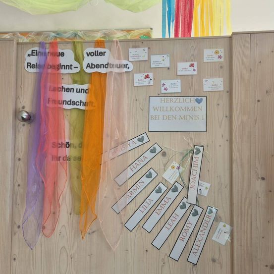 Eine Holztafel zeigt eine bunte Fahne mit Phrasen und Kindernnamen, mit einem Willkommensschild, das 'Herzlich Willkommen bei den Minis 1' lautet.