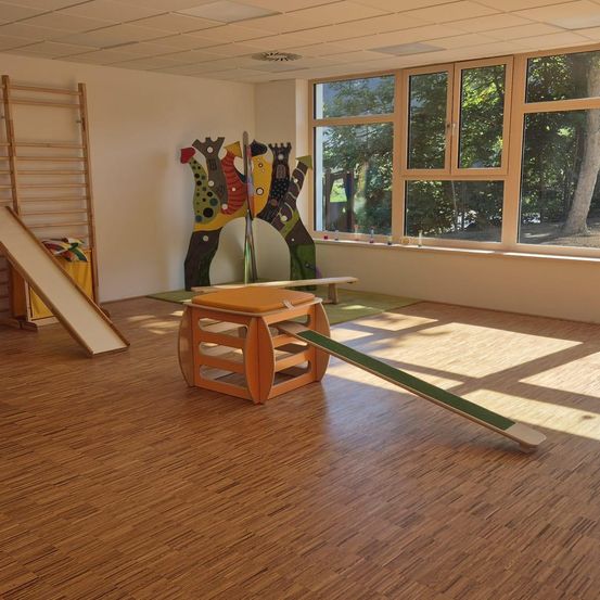 Ein leerer Raum mit Holzböden und großen Fenstern. Ein Rutschen, ein Holzhocker und eine farbenfrohe Wandkunst sind vorhanden. Sonnenlicht strömt durch die Fenster und erhellt den Raum.