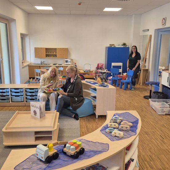 Zwei Frauen in einem Spielzimmer mit Spielzeug, eine Frau sitzt auf einem Kissen, während die andere eine Tablett mit Bällen hält. Es gibt Holzregale, Stühle und Aufbewahrungsboxen. Eine Frau steht in der Nähe einer Tür.