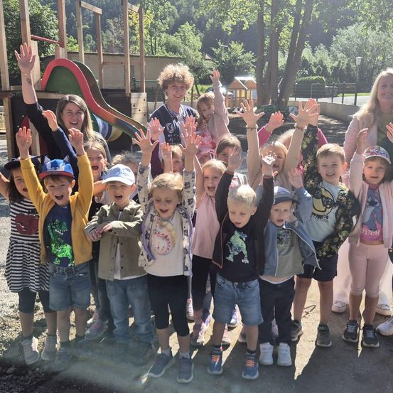 Eine Gruppe junger Kinder und Erwachsener lächelt und winkt zur Kamera in einem Park mit einem Spielplatz im Hintergrund.