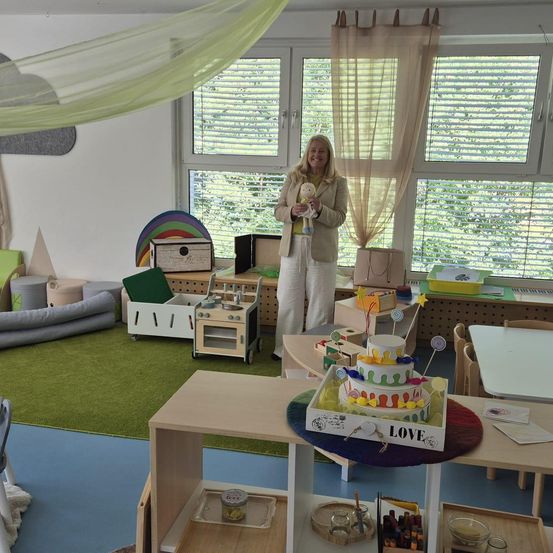 Eine Frau steht in einem Spielzimmer mit Spielzeug und einem Kuchen. Der Raum ist in hellen Farben gehalten und ein großes Fenster zeigt Bäume draußen.