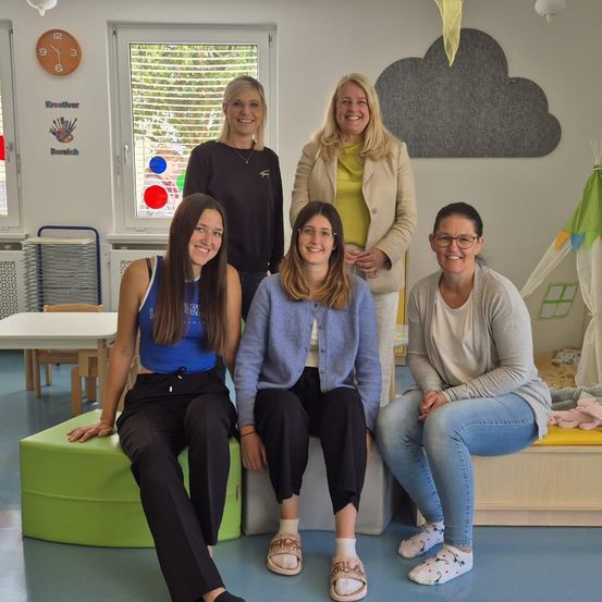 Fünf Frauen sitzen in einem hell erleuchteten Raum. Drei sitzen auf grünen Bänken und zwei stehen dahinter. Der Raum ist mit bunten Akzenten dekoriert und hat ein Spielzelt.