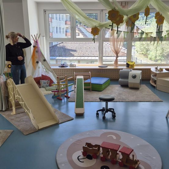 Ein Spielzimmer mit Spielzeug, einer Rutsche und einem Fenster mit Blick auf ein Gebäude. Eine Frau steht neben einem Zelt und einem Stuhl.