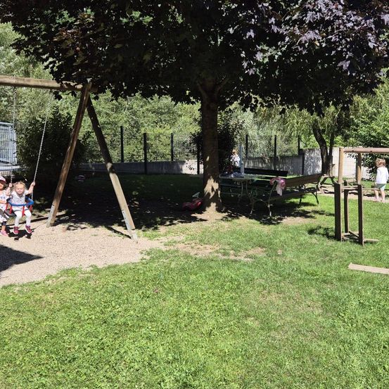 Eine Spielplatzszene mit einer Schaukel und einer Bank unter einem Baum, zwei Kinder schwingen und ein anderes geht in der Nähe. Der Hintergrund zeigt einen Zaun und Grün.