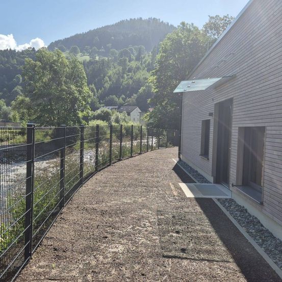 Ein modernes Gebäude mit einer Glasüberdachung steht neben einem Kiesweg mit einem schwarzen Zaun. Ein Fluss und üppige Grünflächen im Hintergrund, mit Bergen und einem Haus sichtbar.