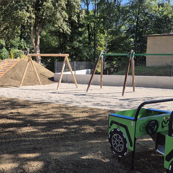 Ein Spielplatz im Freien mit Sandkasten, Schaukeln und Klettergerüst. Ein grünes Spielzeugauto ist in der Nähe geparkt. Bäume und ein Gebäude im Hintergrund.