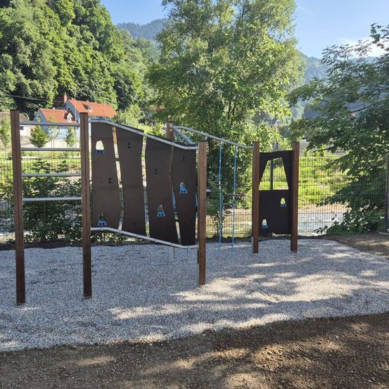 Ein Spielplatz im Freien mit Klettergerüst und Schaukel, umgeben von Bäumen und einem Haus im Hintergrund.