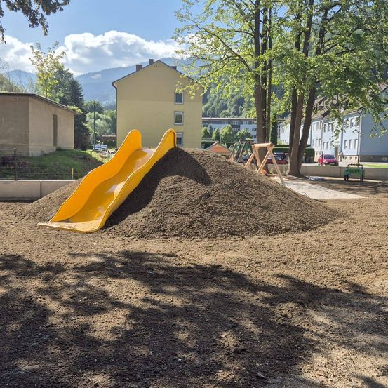 Eine gelbe Rutsche steht auf einem Haufen Erde neben einem Gebäude, mit Bäumen und einem Zaun. Berge sind in der Ferne.