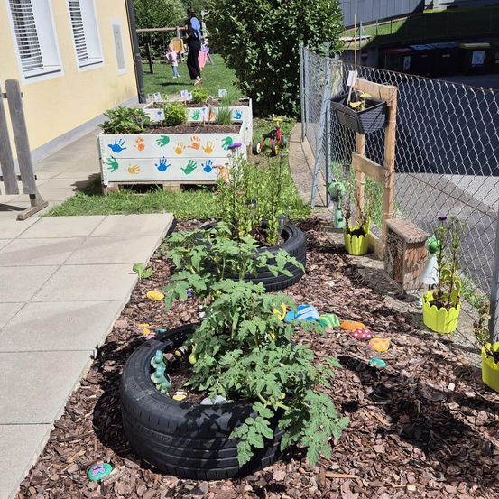 Ein Garten mit Pflanzen in Reifen und Holzkisten mit Handabdrücken steht neben einem Gebäude. Ein Zaun mit einer hängenden Pflanze ist in der Nähe. Eine Person und ein Kind gehen auf dem Rasen.