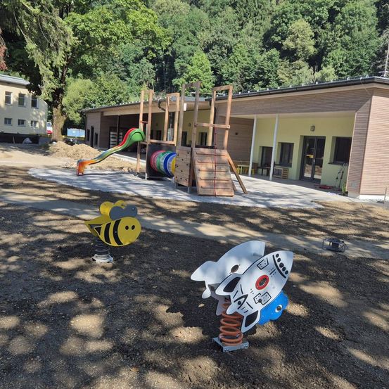 Ein Spielplatz mit einem Bienen-Federreiter, einem Raketen-Federreiter und einer Rutsche vor einem Gebäude mit mehreren Fenstern.