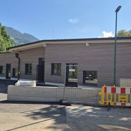 Ein modernes Gebäude mit Holzfassaden und mehreren Fenstern wird gebaut, umgeben von einem Metallzaun und einer Absperrung. Bäume und Berge sind im Hintergrund sichtbar.