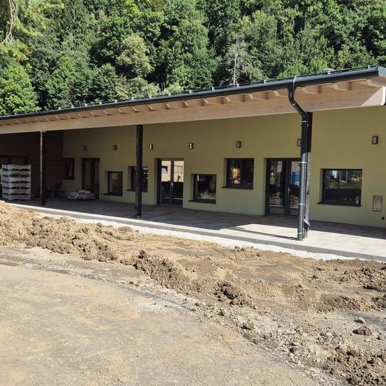 Ein Außenbereich mit einem Gebäude im Bau, das eine Holzfassade, schwarze Metallstützen und mehrere Fenster aufweist. Die Szene umfasst Haufen von Erde, einen Stapel von Kisten und üppige Grünflächen im Hintergrund.