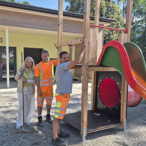 Drei Personen stehen vor einem Spielplatz. Eine Frau hält eine Harke, während zwei Männer neben einem Klettergerüst stehen. Die Umgebung ist im Freien, mit einem Gebäude im Hintergrund.