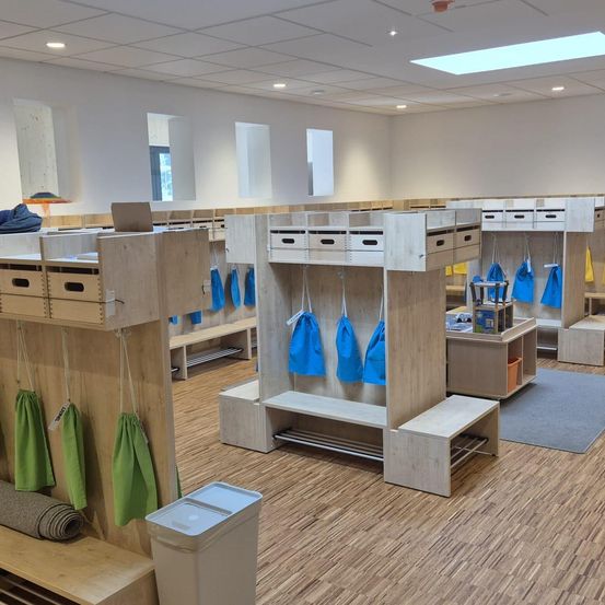 Ein Raum mit Holzlagern und Bänken. Blaue Taschen hängen an Haken, und ein Teppich liegt auf dem Boden. Ein Mülleimer ist in der Nähe.