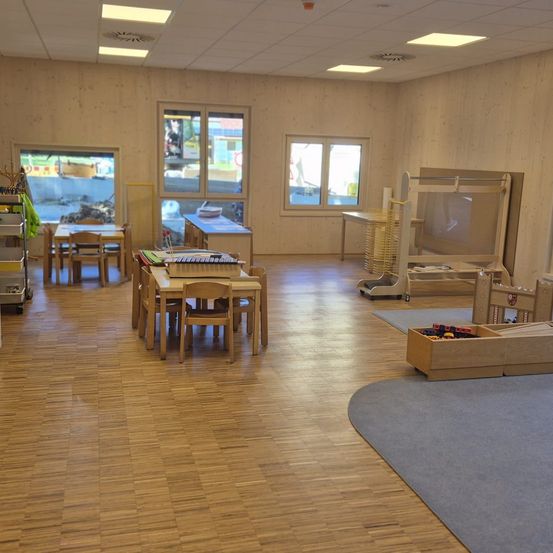 Ein Raum mit Holzböden und -wänden, der Tische, Stühle, Regale mit Gegenständen und einen großen Teppich enthält. Fenster und eine Tür sind sichtbar und bieten Blicke nach draußen.