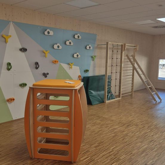 Ein Indoor-Spielbereich mit Holzboden, der eine Kletterwand mit verschiedenen Klettergriffen und eine Holzleiter auf der rechten Seite aufweist. Ein orangefarbener Bau mit Fächern steht vor der Wand.