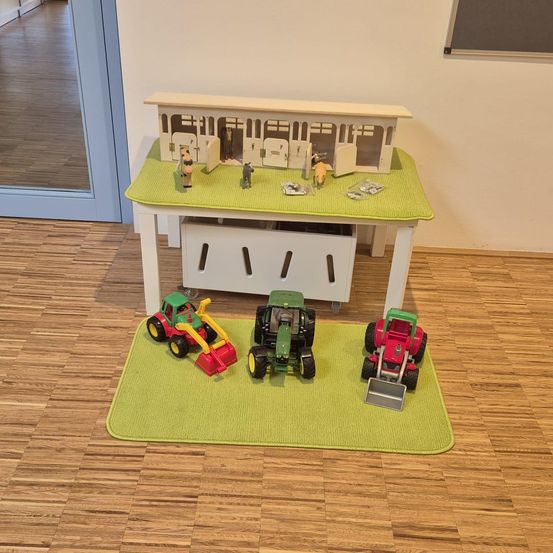 Ein Raum mit einem weißen Schreibtisch und einer grünen Matte, einer Scheune oben mit Pferden und drei Traktoren auf dem Boden. Eine Tür und eine Tafel sind sichtbar.