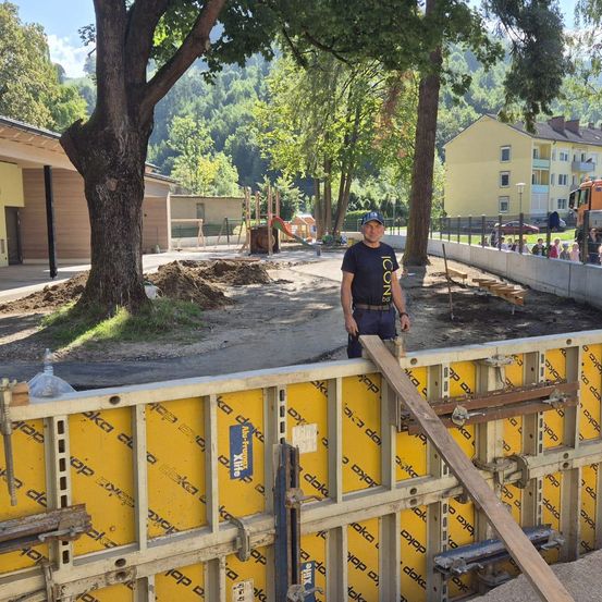 Ein Mann steht neben einer gelben Wand unter einem Baum auf einer Baustelle mit einem Haus und einem Spielplatz im Hintergrund.
