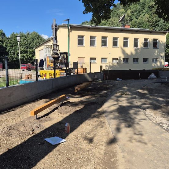 Eine Baustelle mit einem gelben Gebäude, Maschinen und einem Zaun. Ein roter LKW ist draußen geparkt. Bäume sind im Hintergrund.