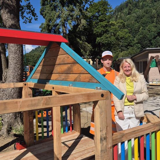 Ein Mann und eine Frau stehen neben einem hölzernen Spielhaus mit rotem Dach und blauer Einfassung. Der Mann trägt ein orangefarbenes Shirt und eine weiße Kappe. Die Frau trägt eine beige Jacke und weiße Hosen. Bäume und ein Haus sind im Hintergrund zu sehen.
