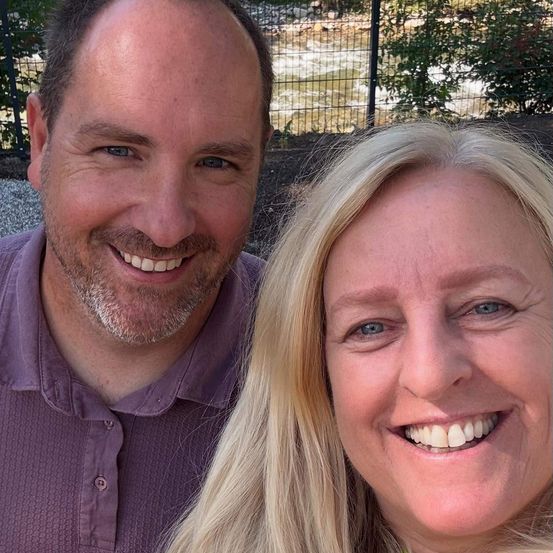 Ein Mann und eine Frau lächeln und posieren für ein Foto, stehen nah beieinander mit einem Zaun und Pflanzen im Hintergrund.