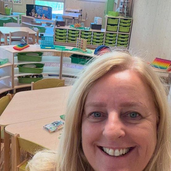 Eine Frau mit blondem Haar lächelt in einem Klassenzimmer mit weißen Regalen, grünen Behältern und einem hölzernen Tisch mit einem bunten Spielzeug.