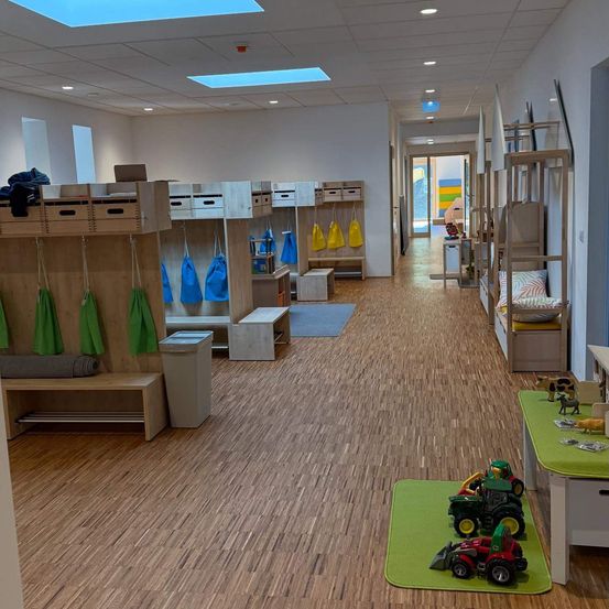 Ein leerer Raum mit Holzböden, Schließfächern und hängenden Taschen. Spielzeug und andere Gegenstände sind auf einem Schreibtisch und einem Teppich platziert.