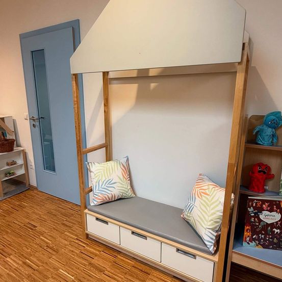 Ein Schlafzimmer mit einem Holzbett mit Kissen, Schubladen und einem Baldachin. Eine blaue Tür und Regale mit Spielzeug sind zu sehen.