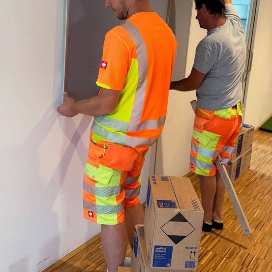 Zwei Männer in orange Sicherheitsanzügen, einer hält ein Brett an eine Wand, der andere mit einem Maßband, in der Nähe von gestapelten Kisten.