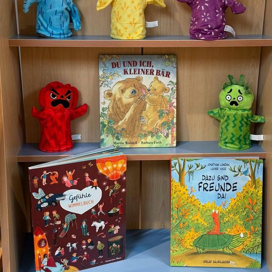 Ein Holzregal mit vier Büchern und vier Plüschtieren darauf. Die Bücher sind mit 'Gefühle Wimmelbuch', 'Dazu sind Freunde da!', 'Du und ich, kleiner Bär' und einem weiteren Buch mit deutschem Text betitelt. Die Plüschtiere sind rot, gelb, lila und grün.